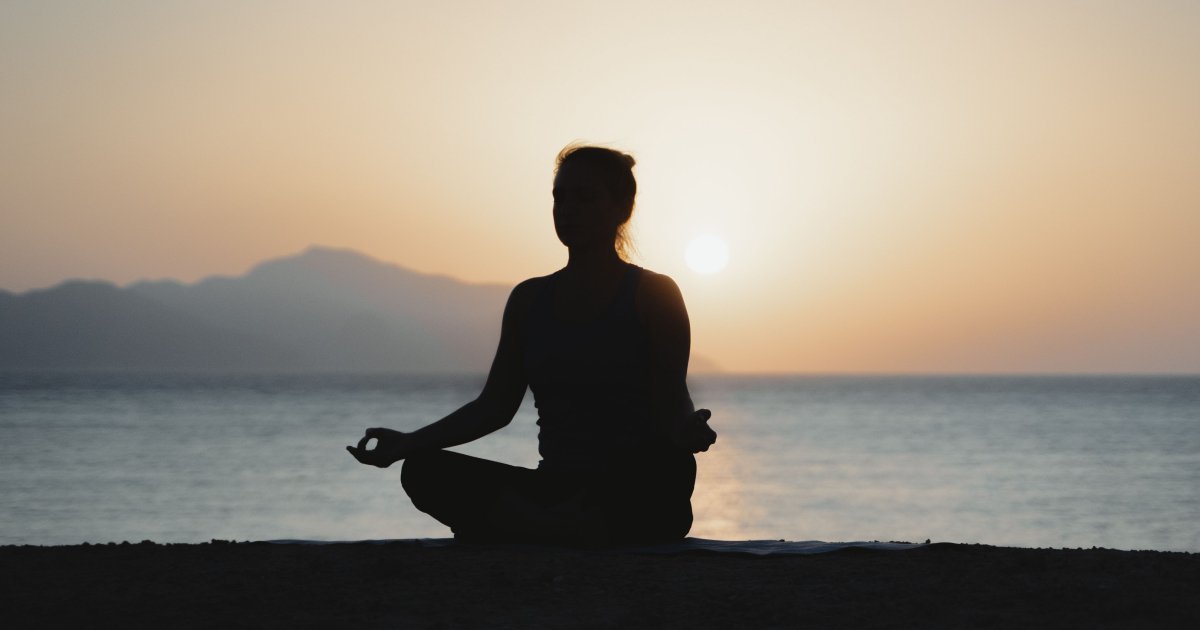 A silhouette of a woman meditating on a beach with the sun setting over her shoulder.